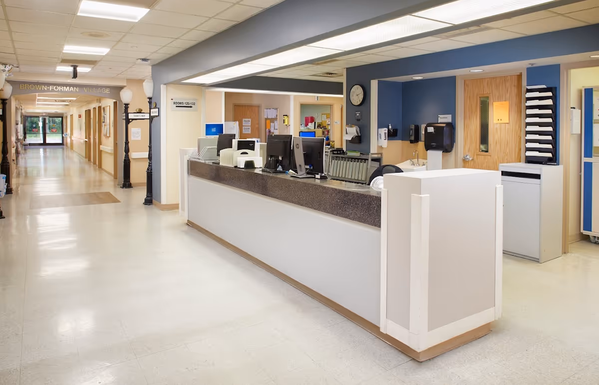 Reception desk area inside Southern Indiana Rehabilitation Hospital - Skilled Nursing Facility with computer monitors, office supplies, and a hallway labeled Brown-Forman Village leading to rooms 120-132.