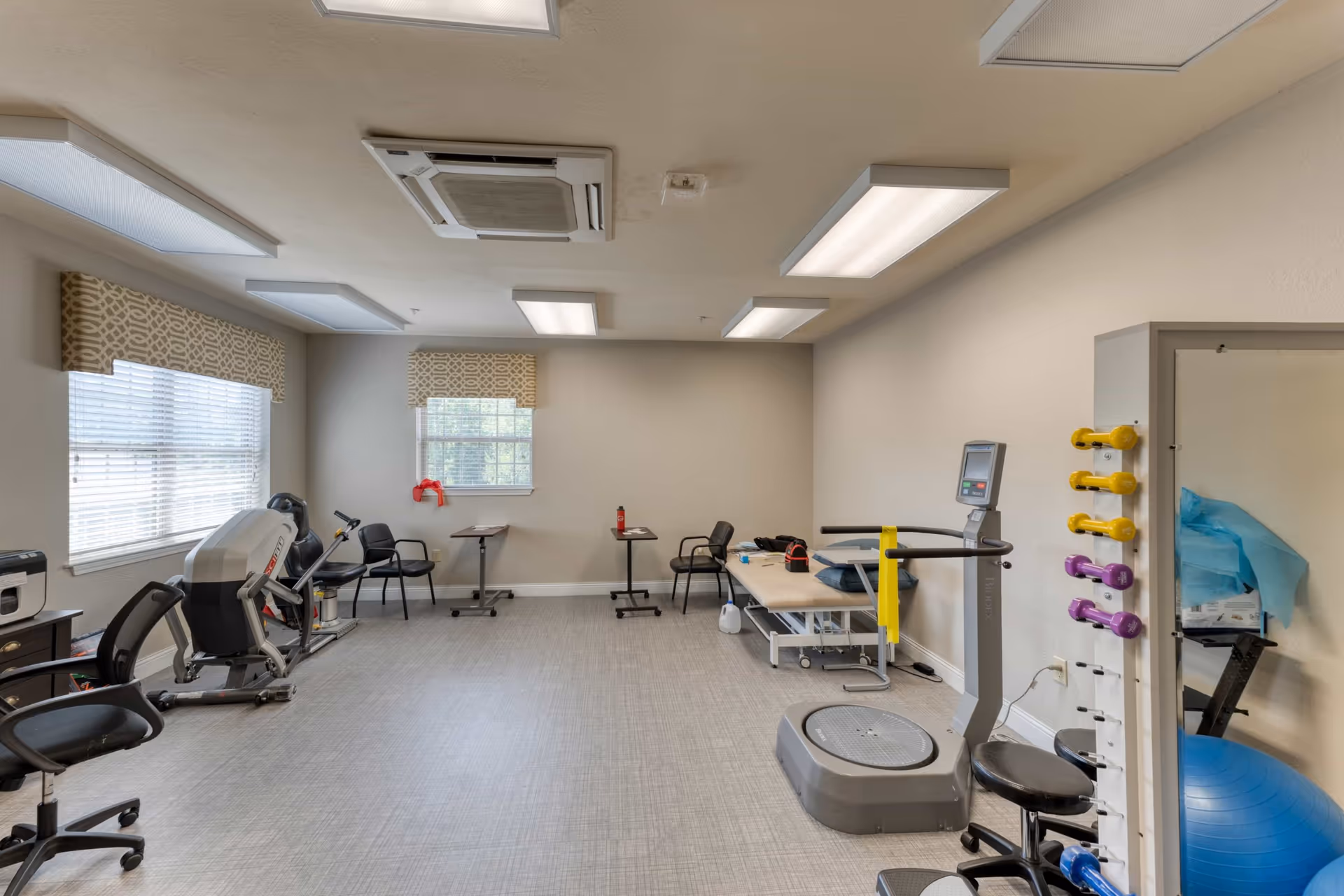 A physical therapy or exercise room with various equipment including a vibration plate machine, small dumbbells on a rack, exercise balls, chairs, and tables. The room has two windows with patterned valances, fluorescent ceiling lights, and a neutral color scheme.