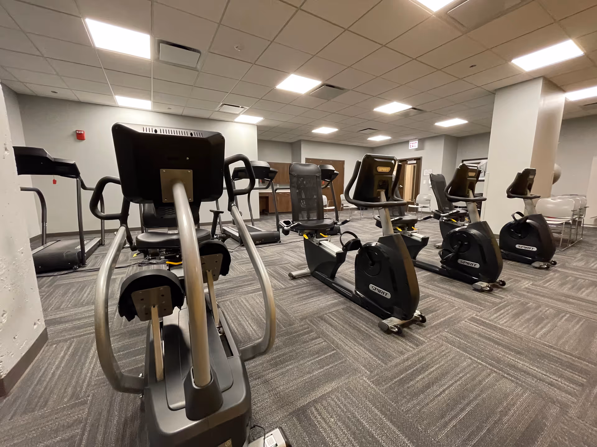Indoor fitness room with several exercise machines including recumbent stationary bikes and treadmills, carpeted floor, and bright overhead lighting.
