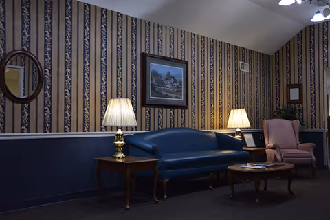 A cozy sitting area with a blue leather sofa, a pink upholstered armchair, two wooden side tables each with a lamp, a round wooden coffee table with magazines, and framed artwork on the wallpapered wall.
