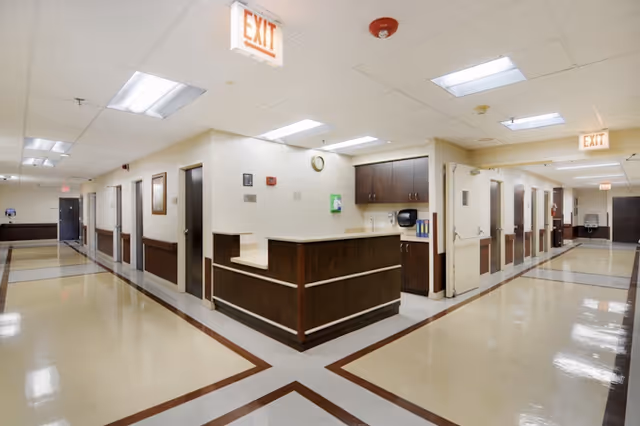Interior view of a clean, well-lit hallway in a senior living facility with a central nurse's station featuring dark wood cabinetry. The hallway has multiple closed doors on both sides, beige walls, and a tiled floor with brown accents. Exit signs and ceiling lights are visible.