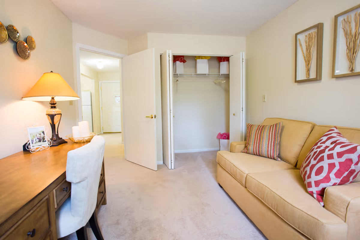 Well-lit living room/den with a beige sofa and patterned pillows, a desk with lamp and chair, and an open closet.