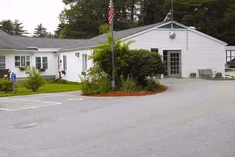 A single-story white senior living center exterior with a driveway, flagpole, and landscaped entrance.