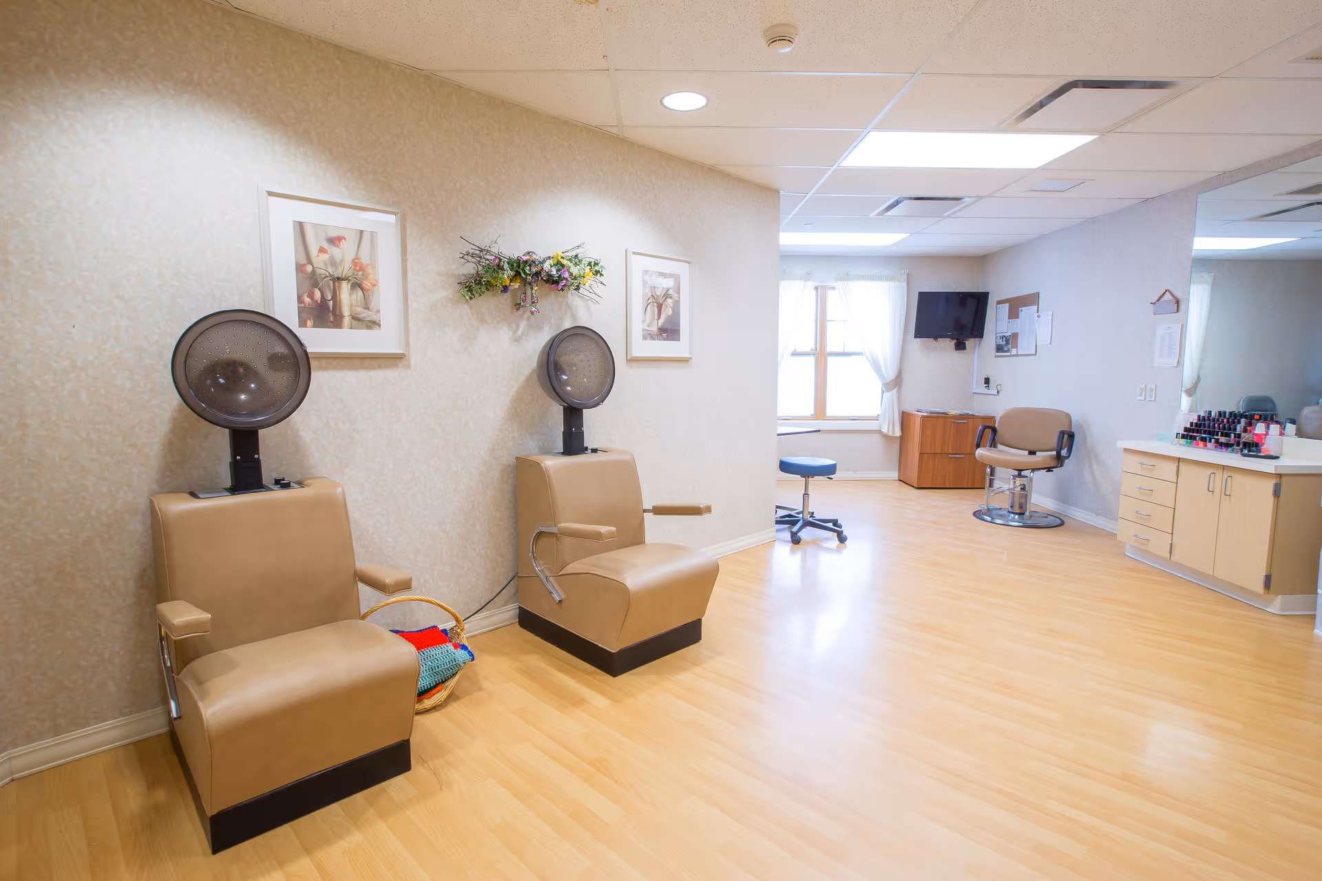 Interior of a hair salon area in a senior living facility with two beige salon chairs equipped with hair dryers, a basket with colorful blankets, framed floral pictures on the wall, a small stool, a TV mounted on the wall, a corkboard, and a counter with various nail polish bottles.