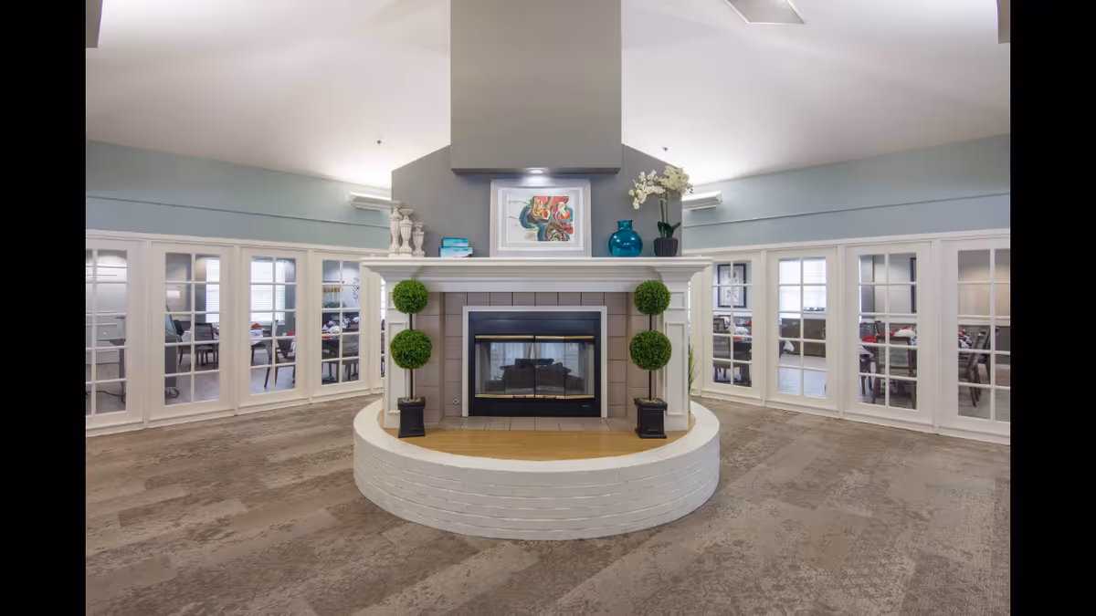 A spacious interior common area featuring a central double-sided fireplace with decorative plants and artwork above it. Surrounding the fireplace are glass-paneled doors leading to rooms with tables and chairs. The room has a high ceiling and neutral-toned carpet flooring.