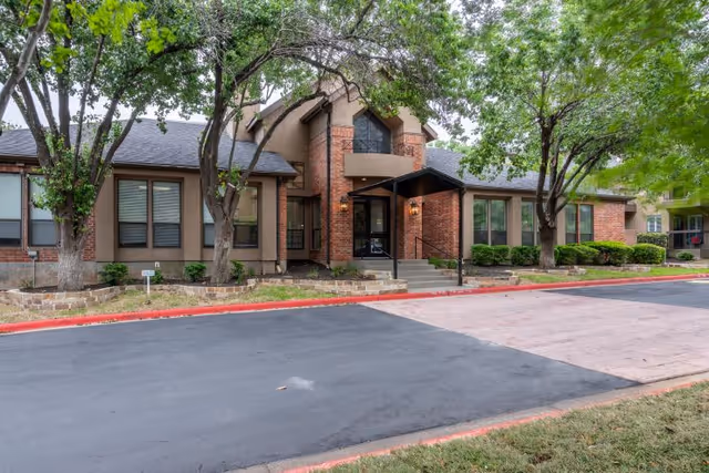 Exterior view of a single-story brick and stucco building with a covered entrance, surrounded by trees and landscaping, with a paved driveway and red curb in front.