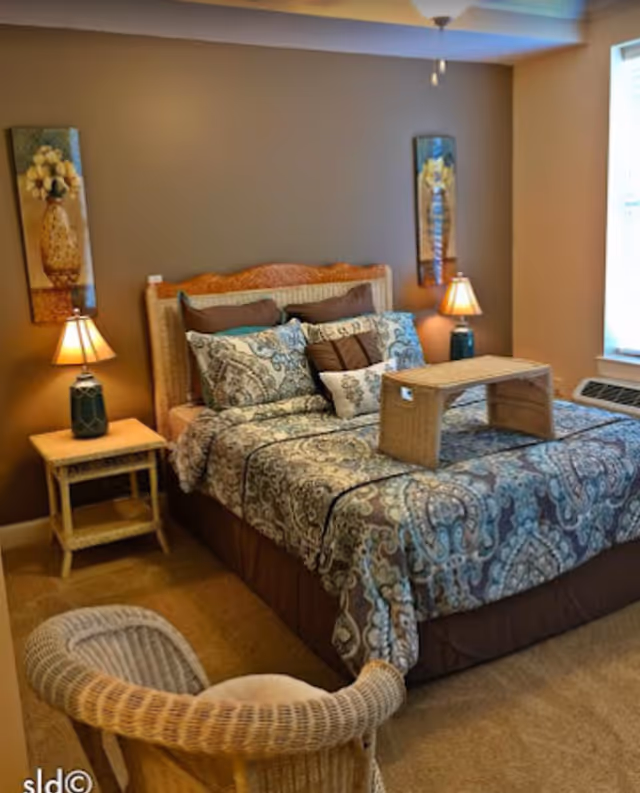 A cozy bedroom with a bed featuring a patterned blue and brown comforter and multiple pillows. There is a wicker tray on the bed. On either side of the bed are matching nightstands with table lamps. Two framed floral paintings hang on the wall above the nightstands. A wicker chair is positioned in the foreground, and a window with blinds is visible on the right side of the room.