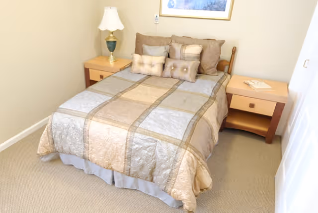 Small bedroom with a double bed dressed in beige and gold bedding flanked by two nightstands and a lamp.