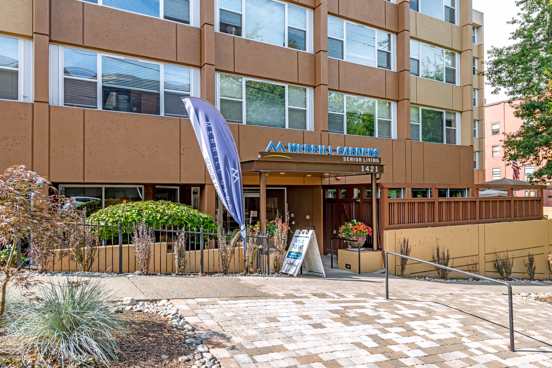 Exterior view of Merrill Gardens Senior Living building entrance with a brown facade, large windows, a blue flag with Merrill Gardens branding, and a signboard near the door. The entrance is surrounded by plants and a paved walkway with a handrail.