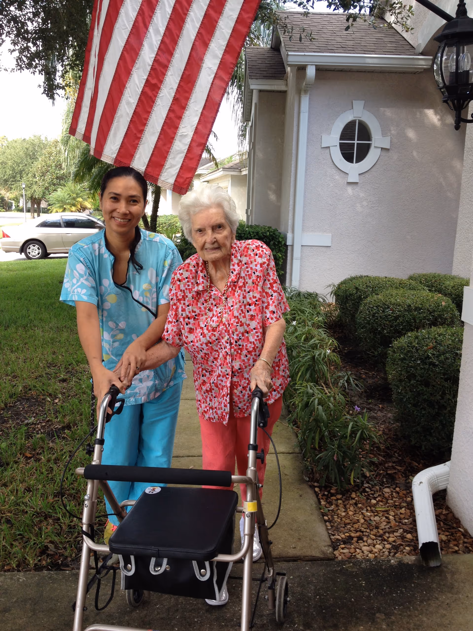 An elderly woman using a walker is standing outside on a sidewalk next to a caregiver in blue scrubs. They are smiling and holding hands. Behind them is a beige building with a round window, some bushes, and an American flag hanging above.
