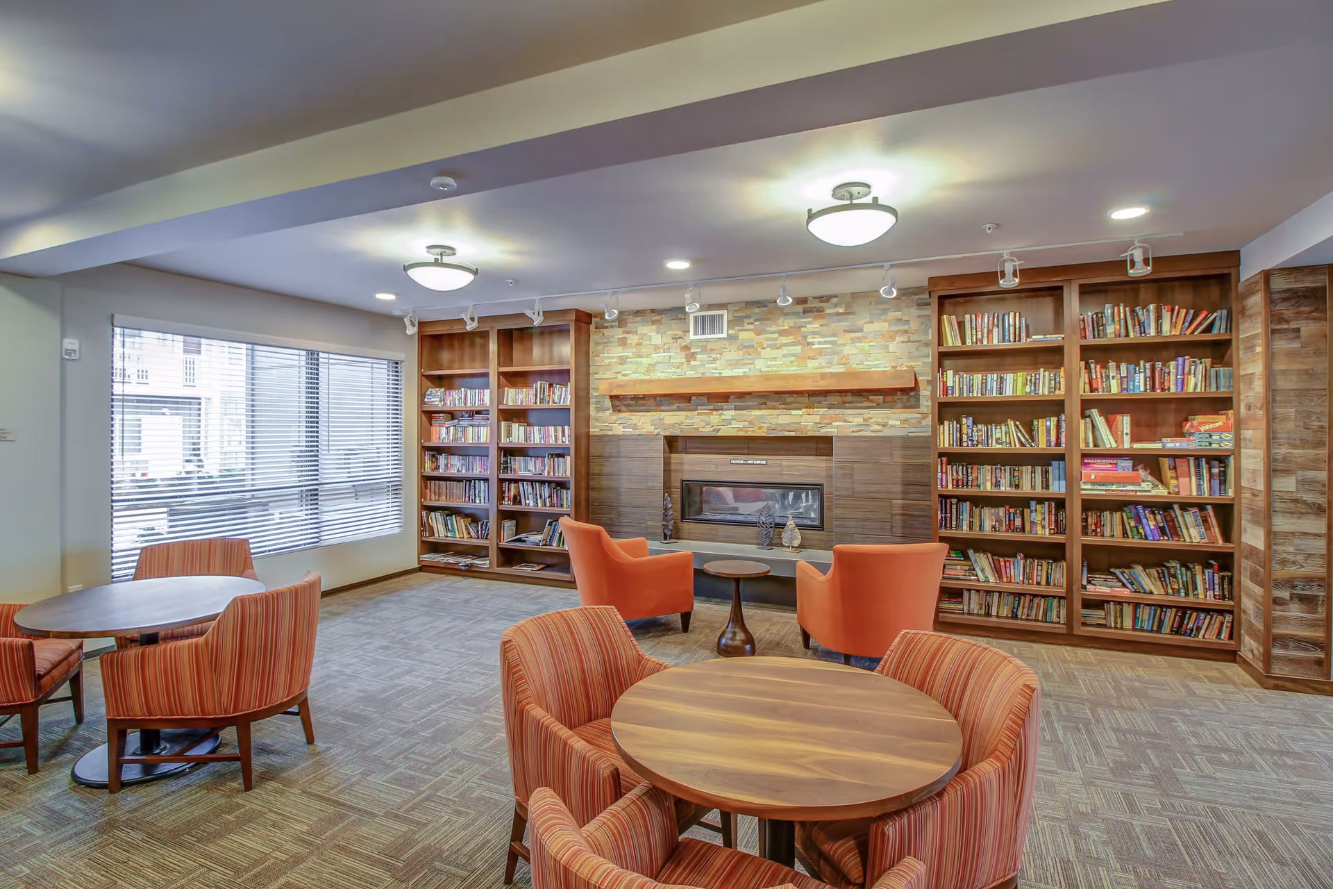 Community lounge with bookshelves flanking a fireplace, round wooden tables and striped armchairs under ceiling lights.