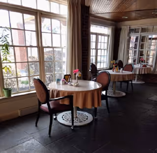 A dining area with round tables covered with beige tablecloths and set with small flower vases. Each table is accompanied by two upholstered chairs with armrests. Large windows with white frames allow natural light to fill the room, and the floor is made of dark tiles. The walls feature brick and wood paneling, creating a warm and inviting atmosphere.