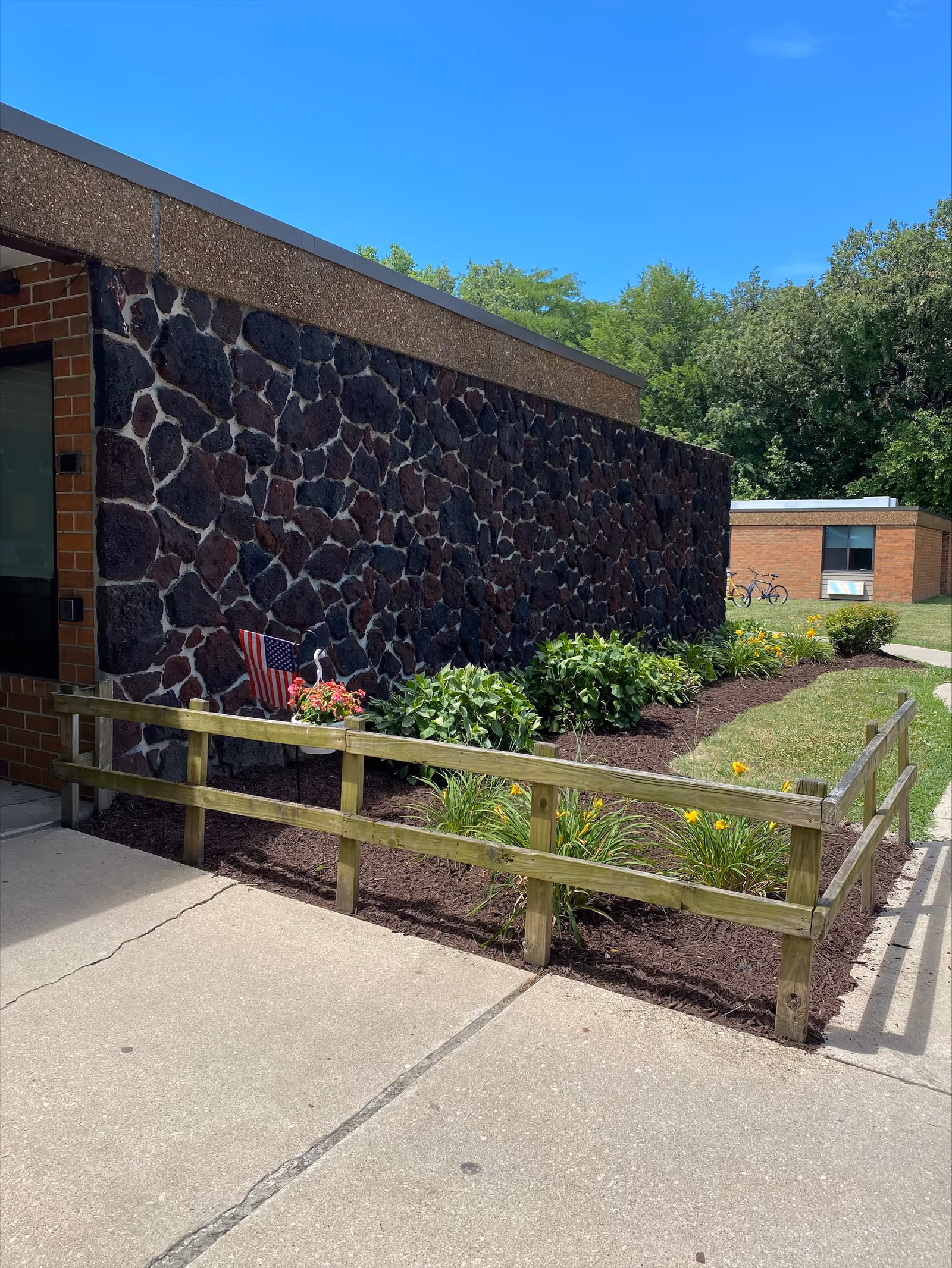 Outdoor view of a building with a stone and brick exterior wall, a small garden bed with green plants and yellow flowers bordered by a wooden fence, and a concrete walkway. In the background, there is another brick building and two bicycles parked on the grass under a clear blue sky.