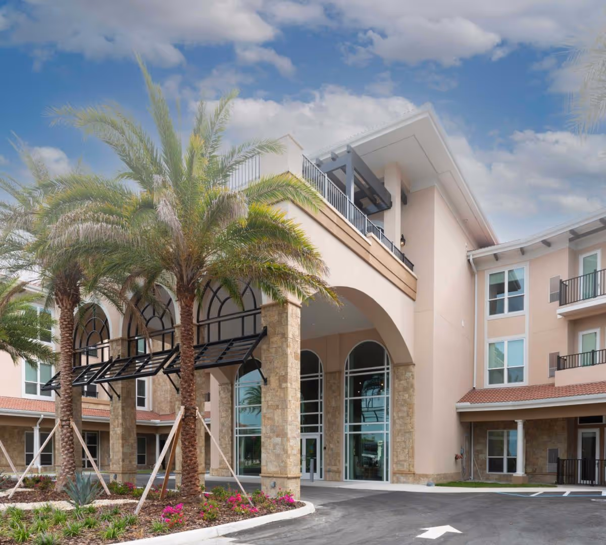 Exterior view of Heartis Venice Assisted Living facility showing a modern building with large arched windows, stone pillars, and palm trees in front under a partly cloudy sky.