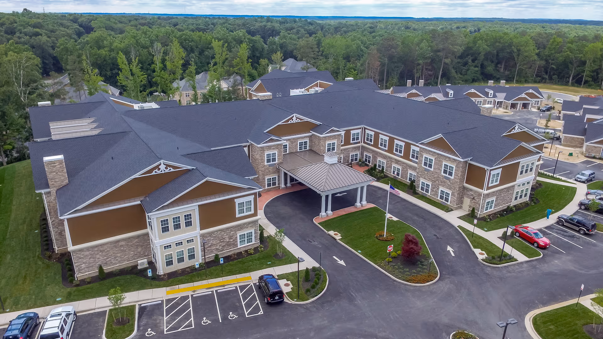 Aerial view of a large assisted living building with a covered entrance, parking lot, and landscaped grounds surrounded by trees.