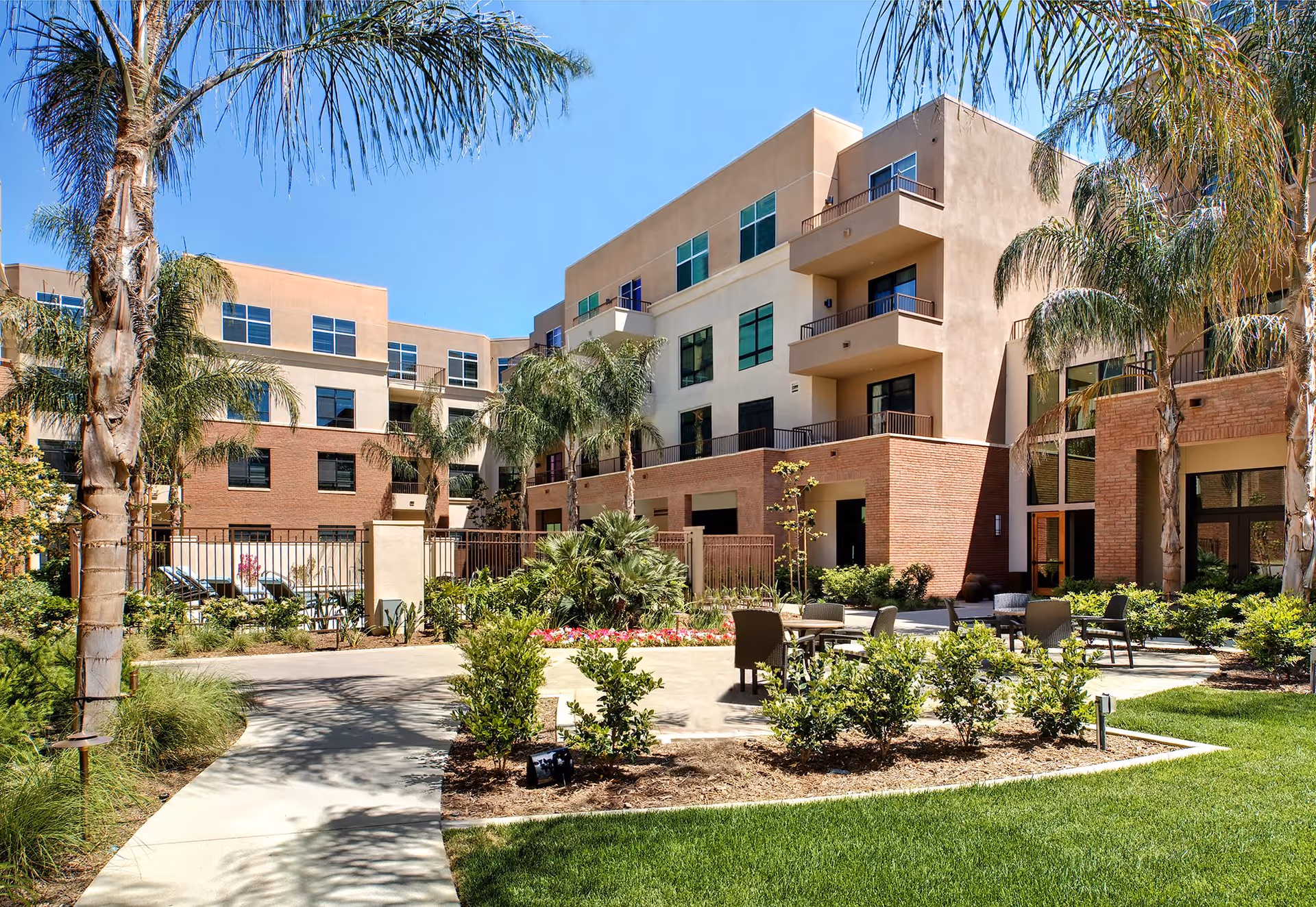 Outdoor courtyard area of The Village at NorthRidge senior living facility featuring a paved walkway, green lawn, palm trees, shrubs, and outdoor seating with chairs and tables. The multi-story building with balconies and large windows surrounds the courtyard under a clear blue sky.