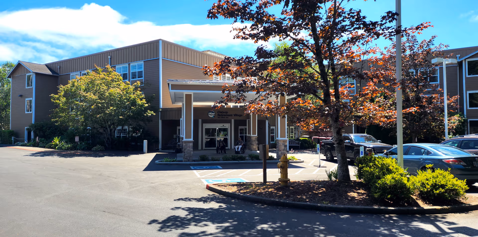 Exterior view of Clatsop Care Center Health District building with a covered entrance, surrounded by trees and parked cars under a blue sky with some clouds.
