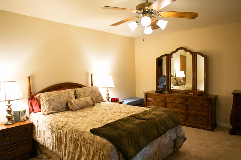 Well-lit bedroom with a made bed, nightstands and lamps, ceiling fan, and a dresser with a mirror.