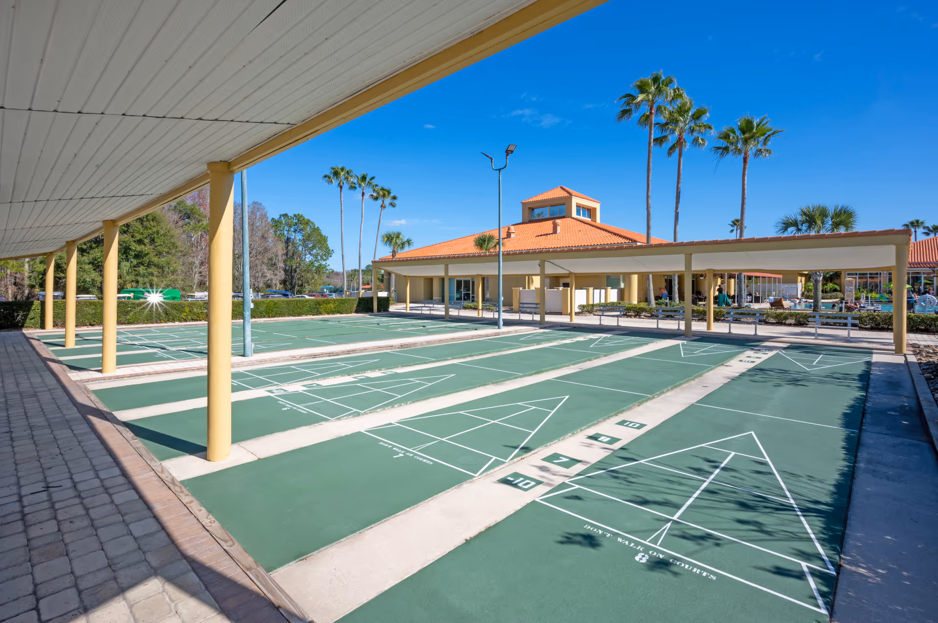 Outdoor shuffleboard courts with green playing surfaces and white markings, covered by a roof supported by yellow pillars. In the background, there is a building with a red-tiled roof and palm trees under a clear blue sky.