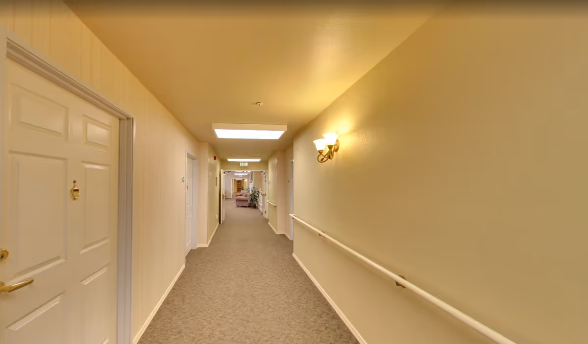 A well-lit hallway in a senior living facility with beige walls, carpeted floor, white doors on the left side, handrails on the right wall, and a light fixture mounted on the right wall. At the end of the hallway, there is a glimpse of a common area with seating and plants.