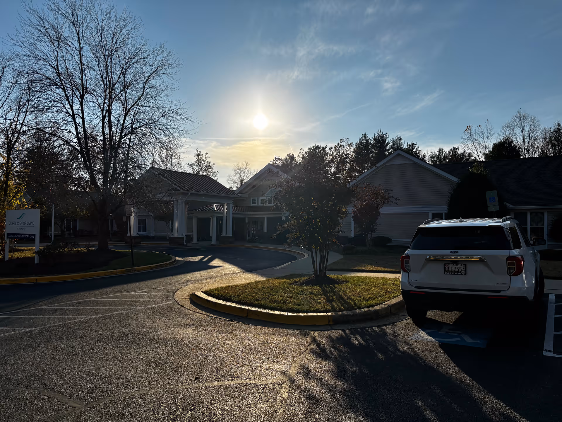 Exterior view of Charter Senior Living Of Bowie facility during sunset with a circular driveway, a white SUV parked in a handicapped parking spot, leafless trees, and a sign indicating assisted living and memory care.