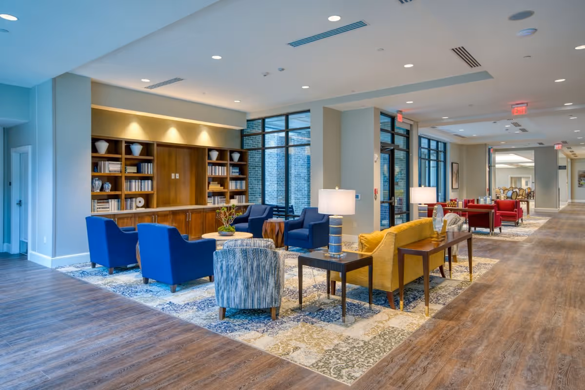 A spacious and well-lit senior living facility common area with a variety of seating options including blue armchairs, a yellow sofa, and patterned chairs arranged around coffee tables. The room features large windows, wooden bookshelves with decorative items, and lamps on side tables. The floor is a combination of wood and patterned rugs, and the space extends into a hallway with more seating and dining areas visible in the background.