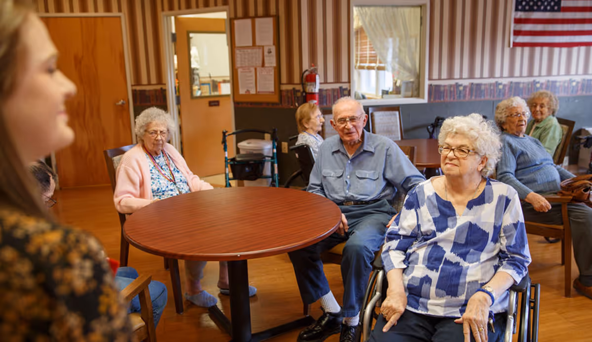 Residents sitting around a round table in a communal room with an American flag on the wall.