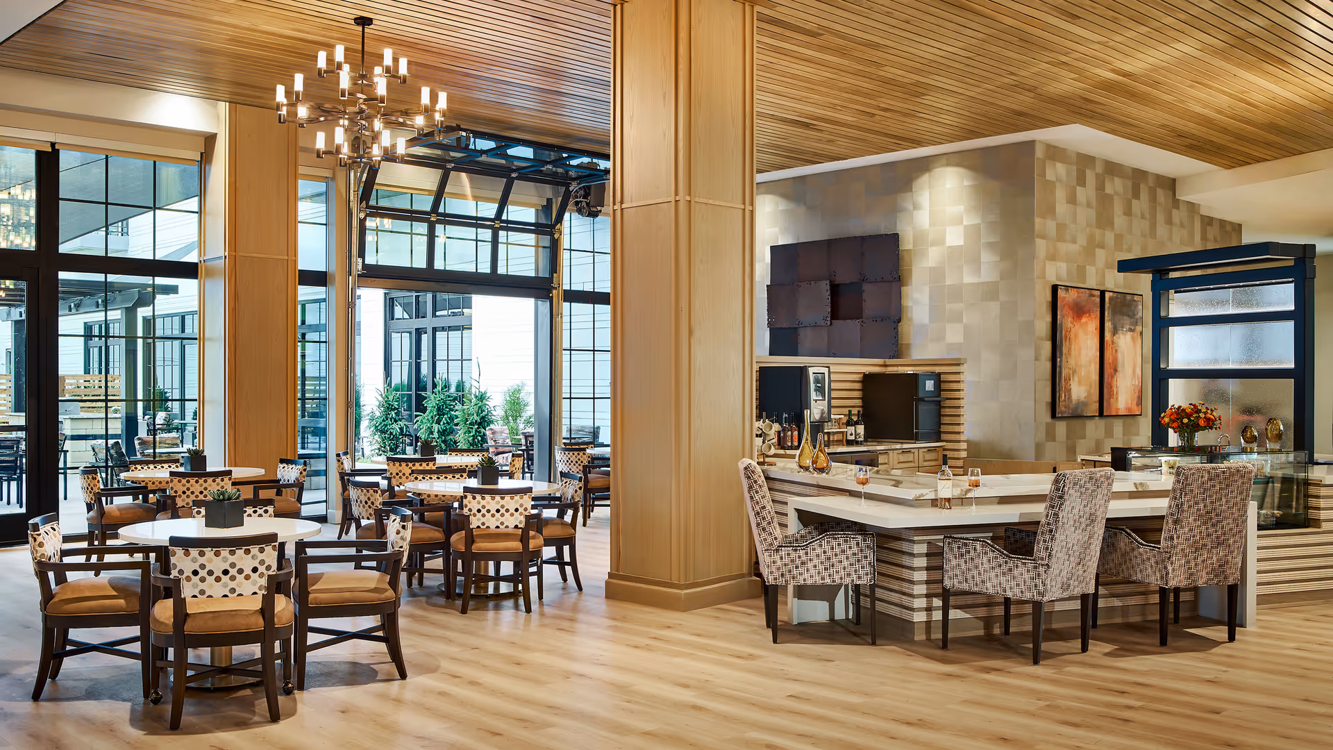 A modern dining area in a senior living facility featuring round tables with cushioned chairs, a wooden ceiling, large windows with black frames, and a bar counter with high chairs. The space is well-lit with a chandelier and natural light coming through the windows, and there are decorative plants and artwork on the walls.