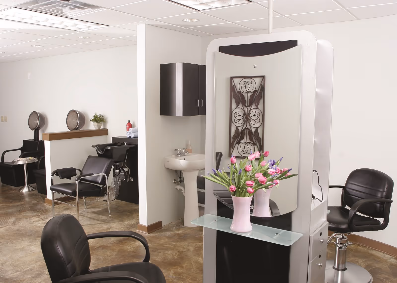 Interior view of a salon area with black salon chairs, hair drying stations, a sink, a large mirror with a vase of pink and purple flowers, and a decorative wall piece.