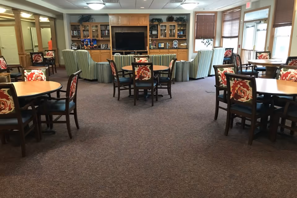 A spacious common area with multiple round wooden tables and chairs featuring floral upholstery. In the background, there are several striped armchairs arranged around a large flat-screen TV set within wooden cabinetry filled with books and decorative items. Large windows with blinds allow natural light into the room.