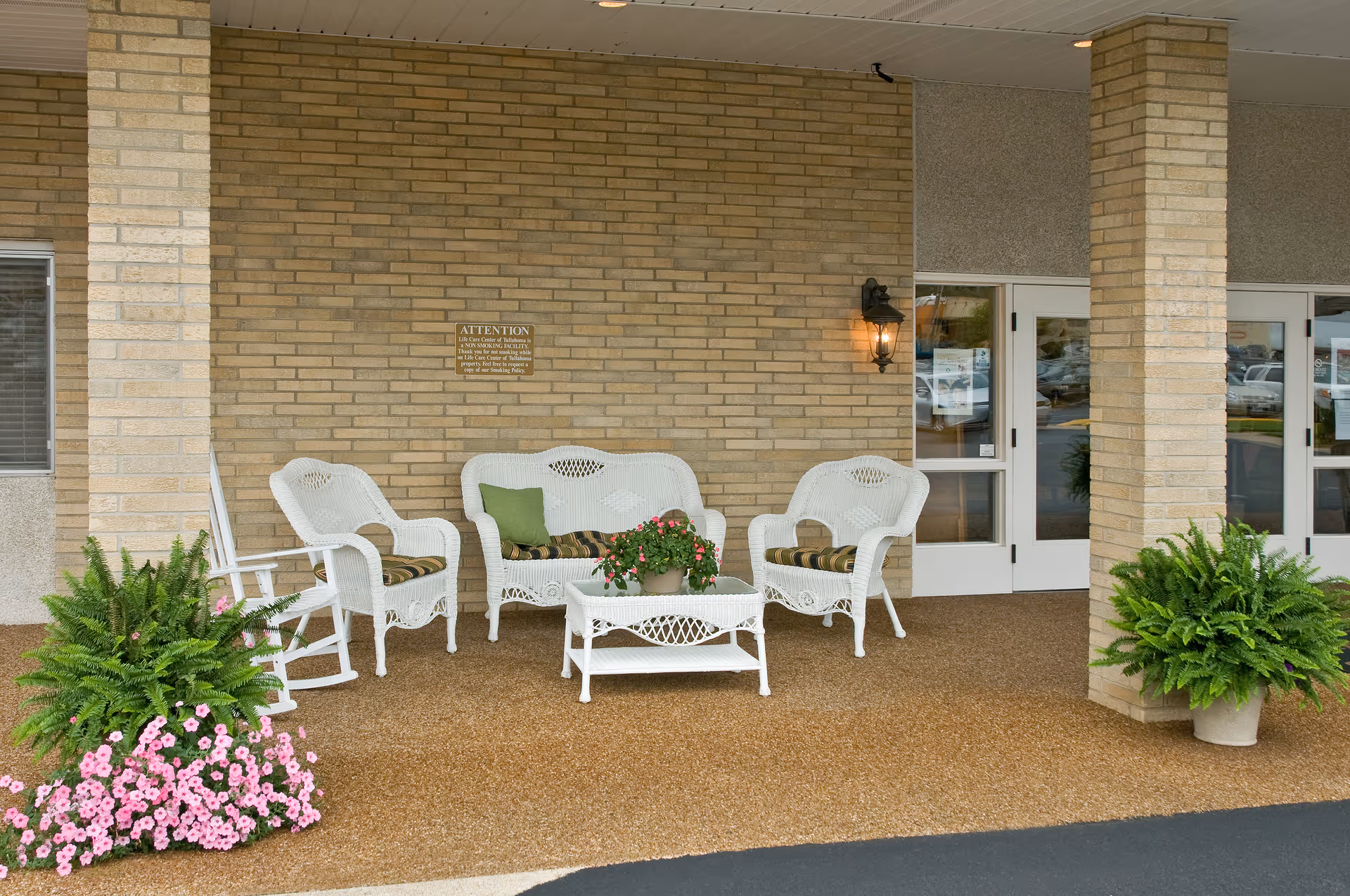 Covered building entrance with white wicker chairs and table, potted ferns and flowers, and brick columns.
