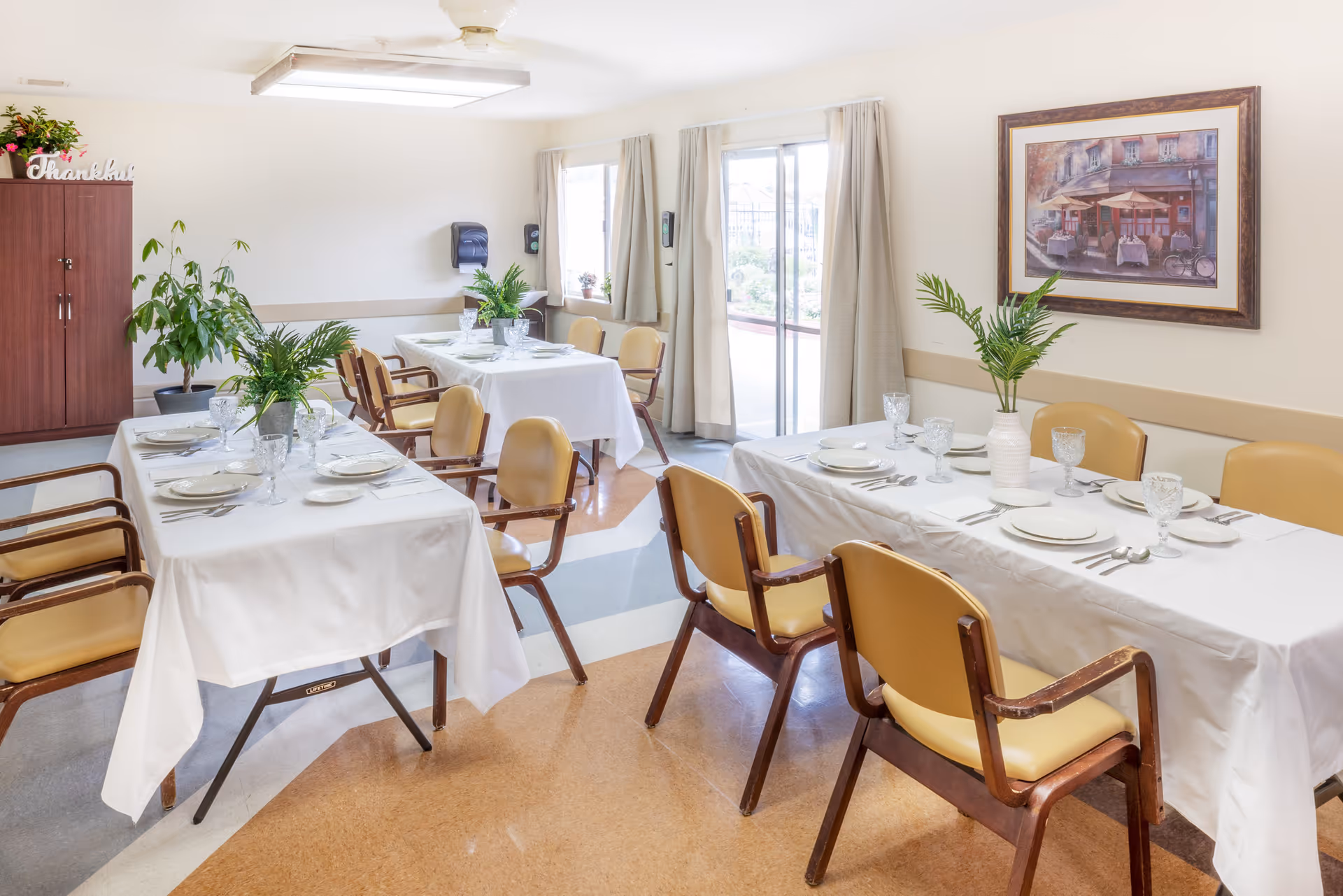Dining room with several tables set for a meal, chairs, potted plants and a framed painting on the wall.