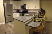 Modern kitchen area with granite countertops, stainless steel refrigerator, stove, microwave, and sink. There are three wooden bar stools at the counter, each set with a green plate and napkin. The kitchen has light-colored cabinets and wood flooring.