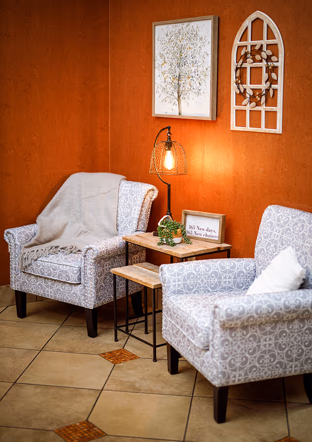 Two patterned armchairs with a small wooden table between them, decorated with a lamp, a small plant, and a framed sign that reads '365 New days, 365 New chances'. The chairs are set against an orange textured wall with two pieces of wall decor: a framed tree artwork and a white windowpane-style decoration with a wreath.