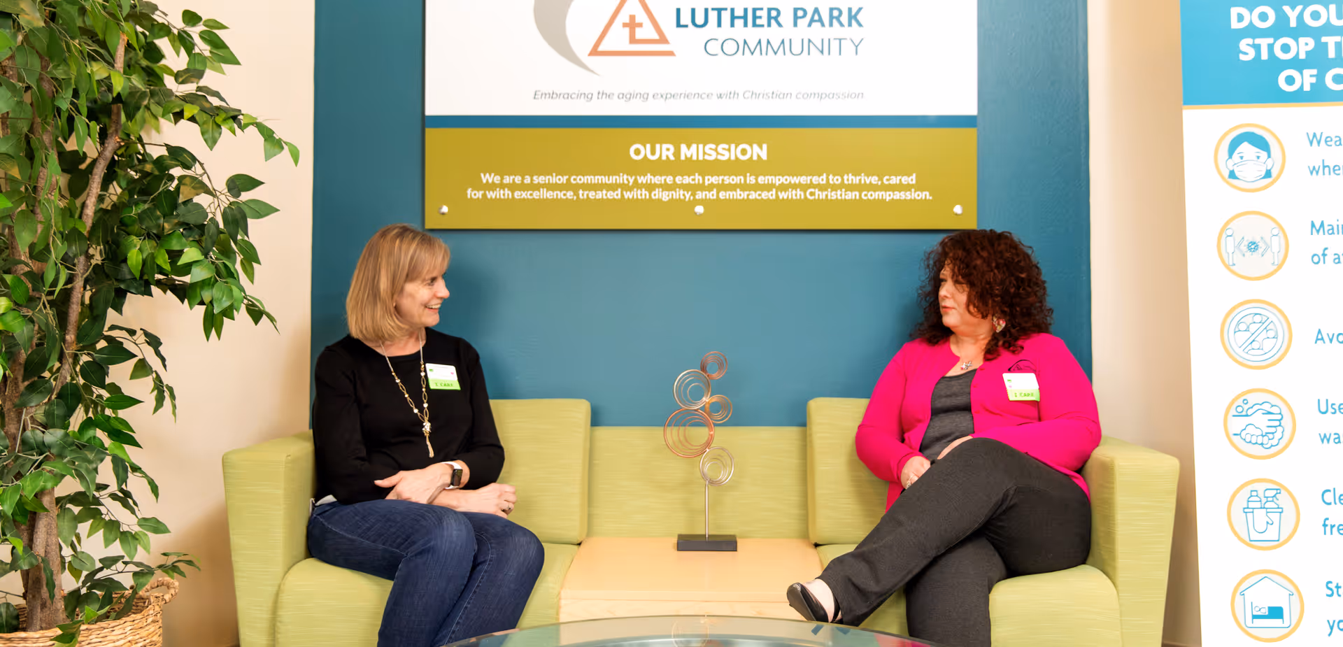 Two women sitting on a light green couch in a waiting area or lounge, facing each other and engaged in conversation. Behind them is a sign with the Luther Park Community logo and mission statement. There is a potted plant to the left and a poster with health guidelines to the right.