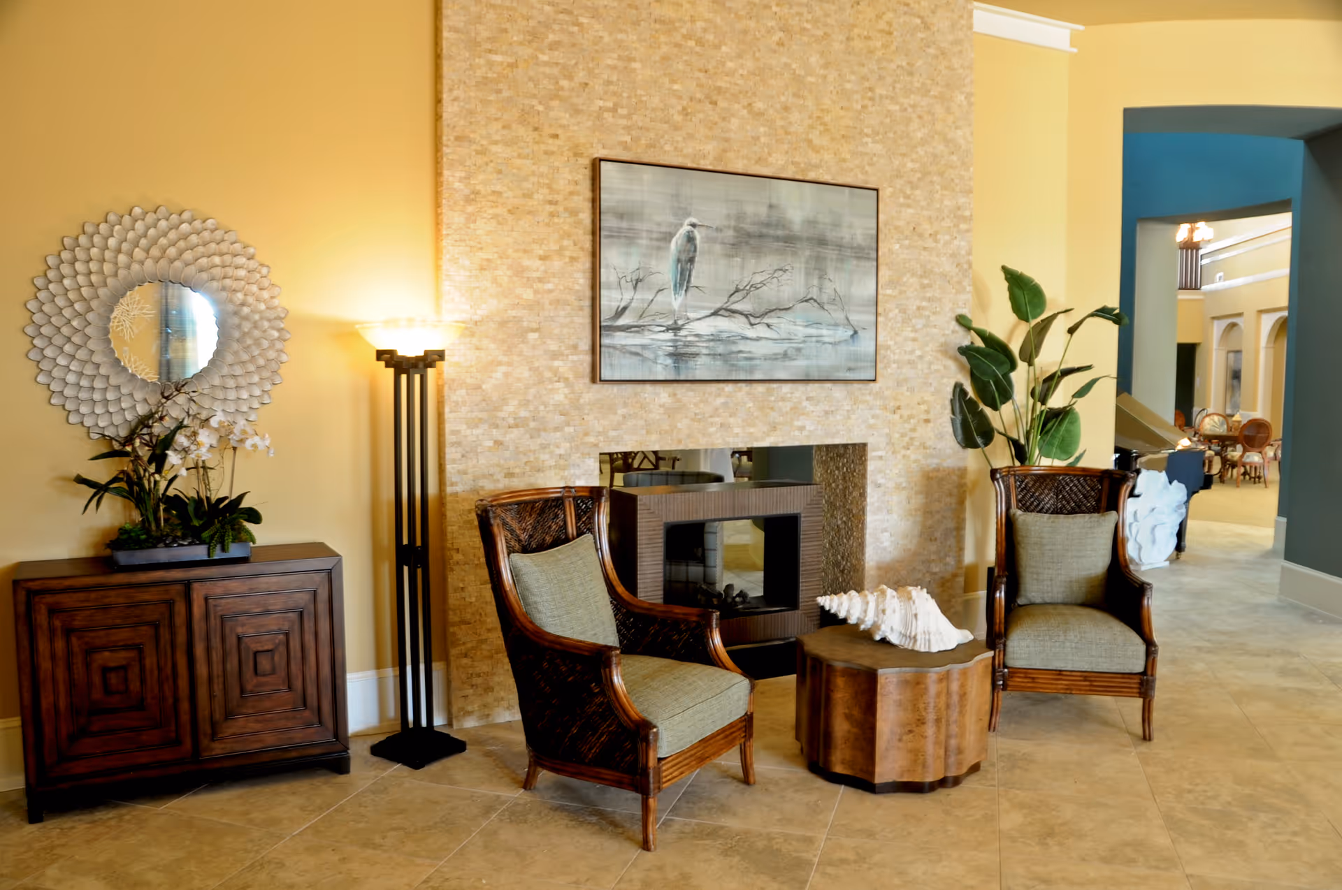 A cozy seating area in a senior living facility with two wicker armchairs with cushions, a small wooden table with a large white seashell on it, a modern fireplace with a stone tile surround, a tall floor lamp, a decorative round mirror above a wooden cabinet with plants, and a framed painting of a bird on a branch above the fireplace.