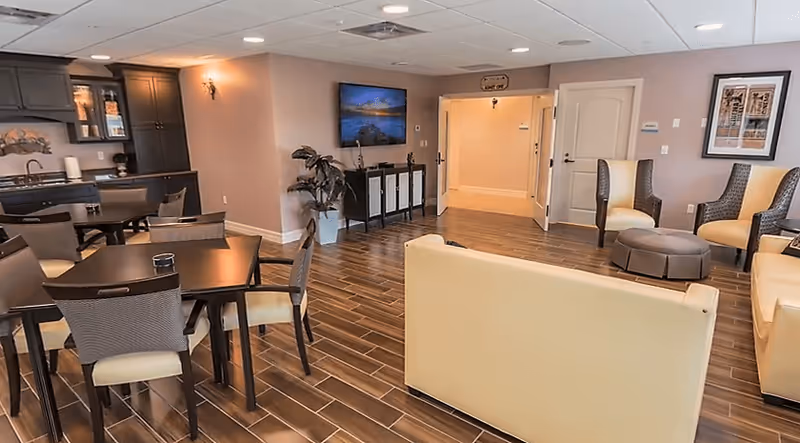 Bright communal lounge and dining area with tables and chairs, sofas and armchairs, a wall-mounted TV, and a small kitchenette.