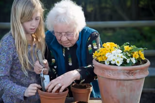 An elderly woman and a young girl are planting flowers together outdoors. The elderly woman is helping the girl place soil into a small flower pot, while a larger pot with yellow and white flowers is nearby.