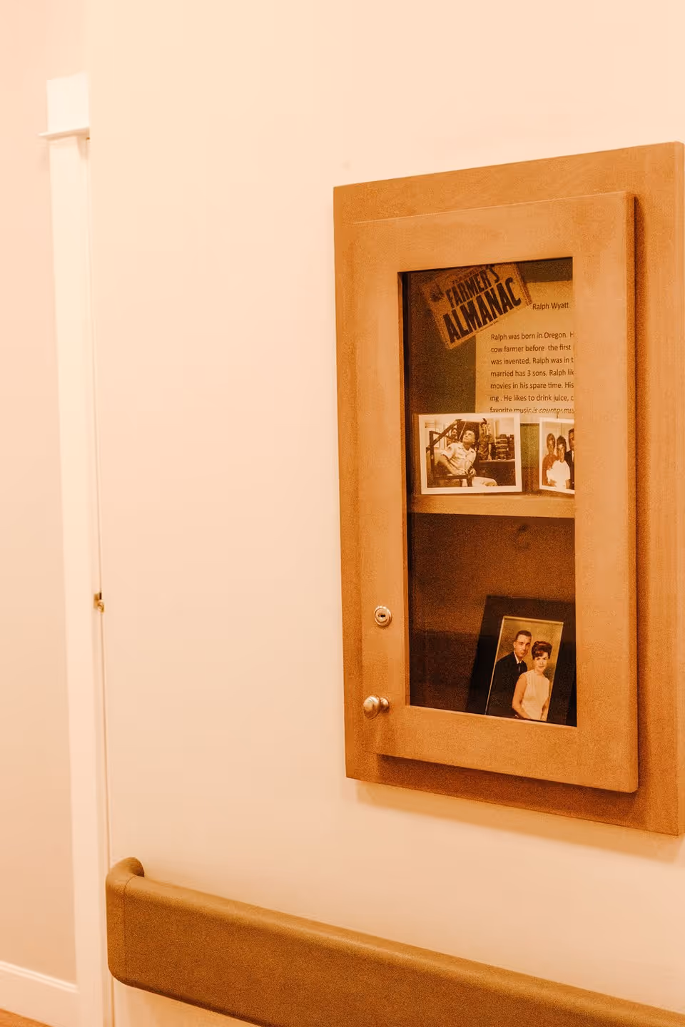 A wooden display case mounted on a beige wall containing old photographs and a printed sheet titled 'The Farmer's Almanac'. Below the display case is a padded handrail attached to the wall.