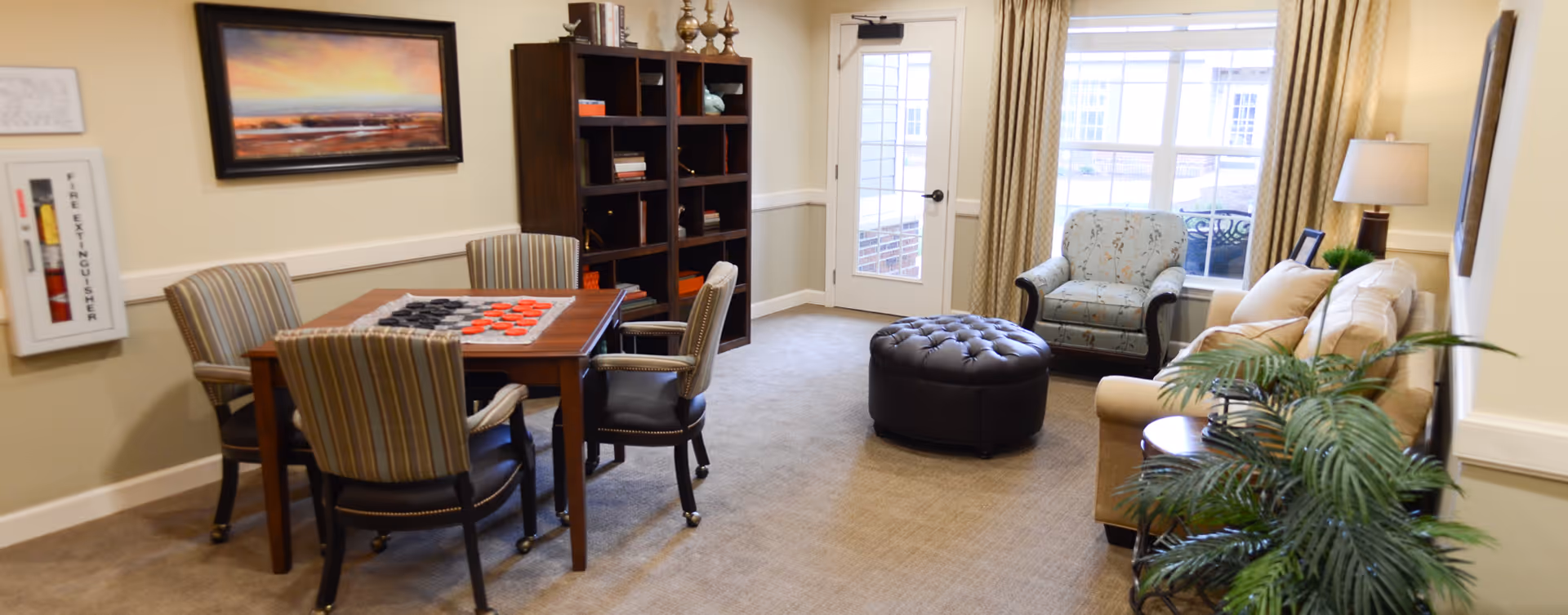 A cozy living room area with a beige sofa, a patterned armchair, a round black ottoman, and a wooden table with four striped chairs arranged around it. The table has a checkers game set up. There is a dark wooden bookshelf with decorative items and books, a lamp on a side table, a large window with curtains, and a door leading outside. A fire extinguisher is mounted on the wall, and a framed landscape painting hangs above the table.