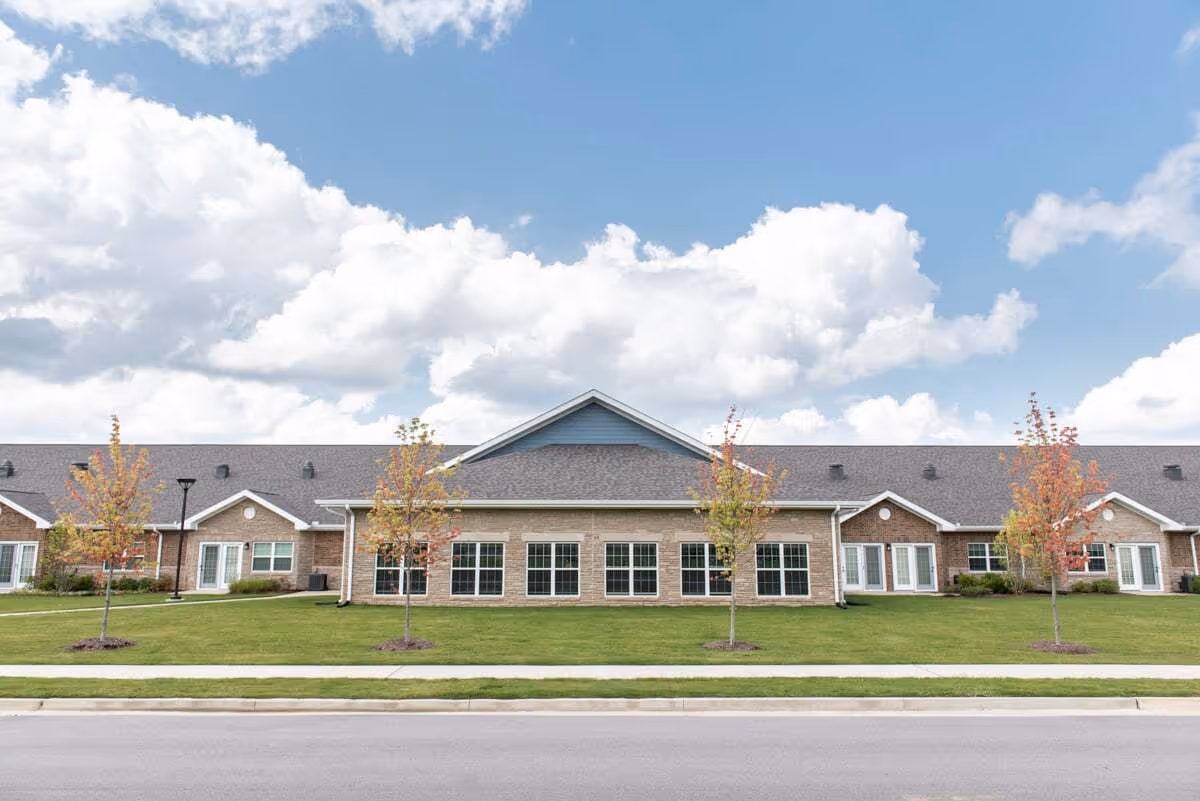 Front view of a single-story brick senior living building with a manicured lawn, small trees, and a cloudy blue sky.