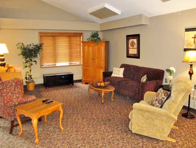 A cozy living room with a patterned carpet, a maroon sofa with decorative pillows, a beige recliner chair, a red patterned armchair, a wooden coffee table with a small centerpiece, a wooden side table with two remote controls, a wooden cabinet, a black TV stand, a potted plant, a floor lamp, and framed artwork on the walls.