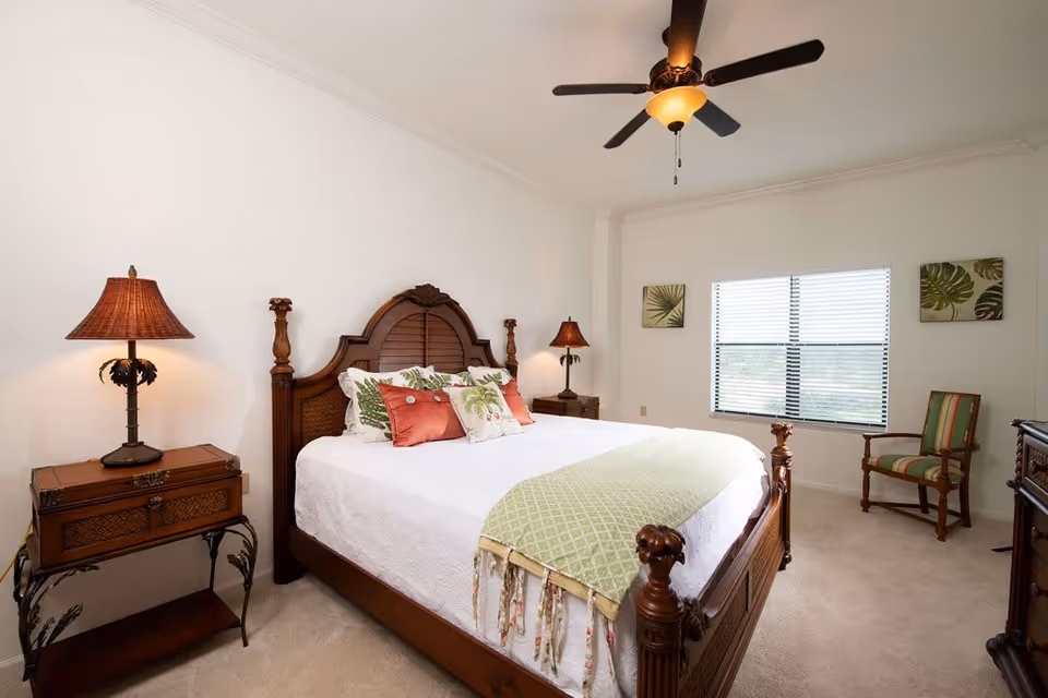 A bright bedroom with a large wooden bed featuring an ornate headboard and footboard. The bed is made with white bedding, green and red decorative pillows, and a green throw blanket. There are two wooden nightstands with matching lamps on either side of the bed. A ceiling fan with a light fixture is mounted on the ceiling. The room has a window with blinds, two framed botanical prints on the wall, and a wooden chair with green and red striped upholstery.