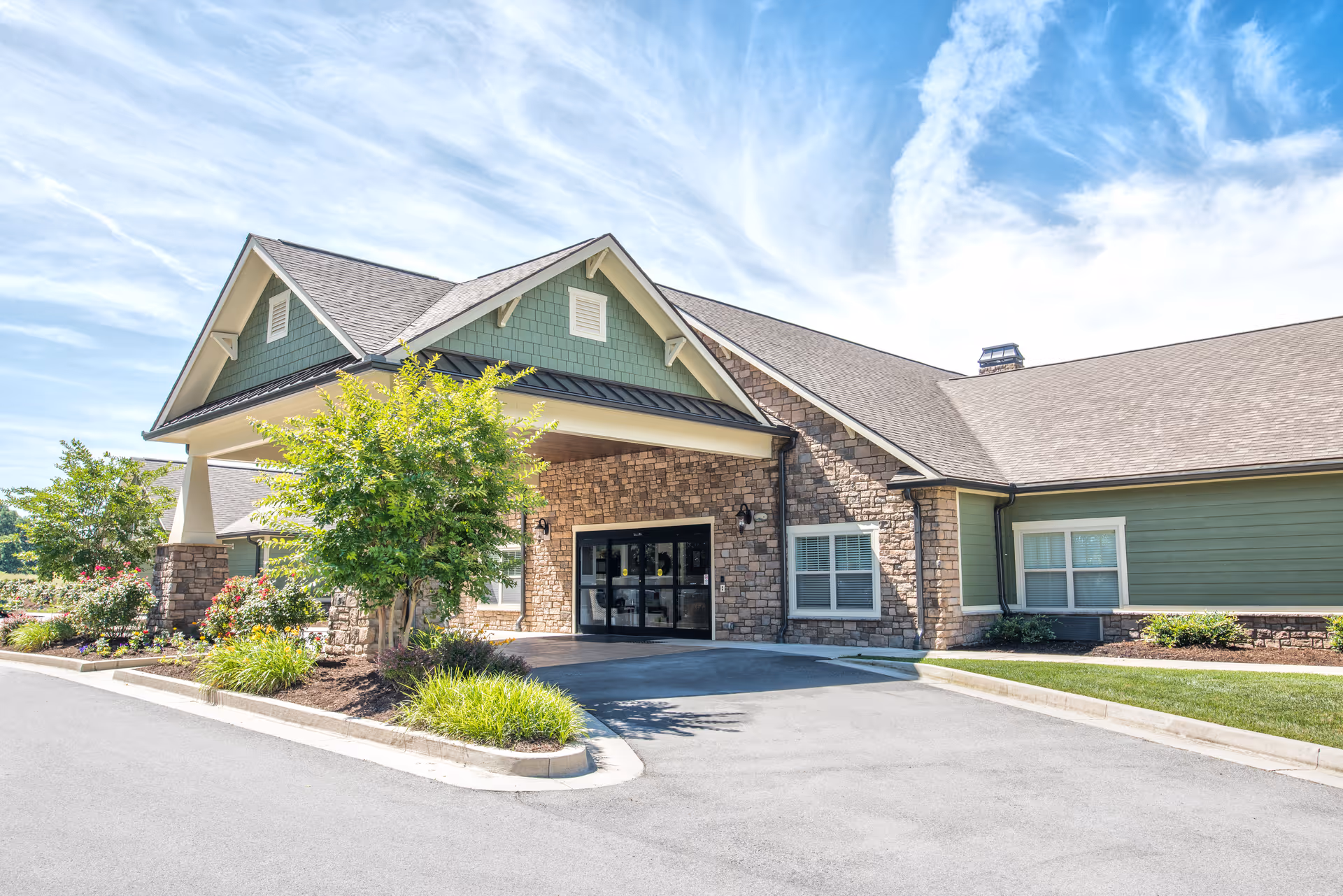 Exterior view of Dominion Senior Living of Crossville showing the entrance with a covered driveway, stone and green siding walls, windows, and landscaped greenery under a partly cloudy blue sky.