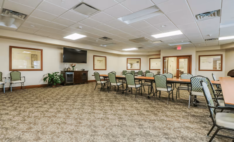 A spacious room with multiple tables and chairs arranged in rows, patterned carpet flooring, beige walls with framed windows, a flat-screen TV mounted on the wall, and ceiling lights providing bright illumination.