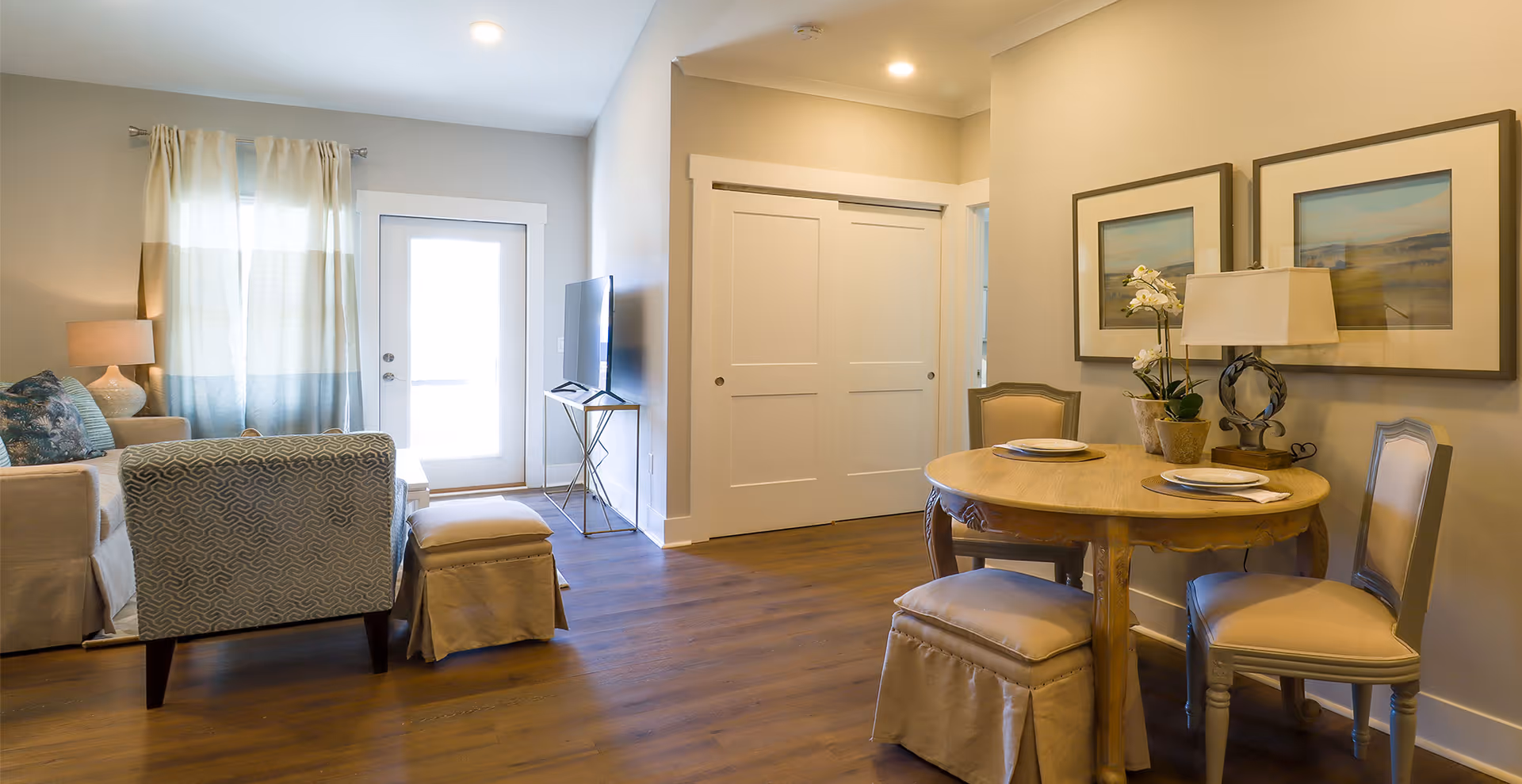 A cozy living and dining area in a senior living facility with wooden flooring. The living area has a sofa, an armchair, an ottoman, a side table with a lamp, and a TV on a stand near a door with a window. The dining area features a round wooden table set for two with plates and napkins, two chairs, and a cushioned stool. The walls are decorated with two framed landscape paintings and a table lamp with a floral arrangement.