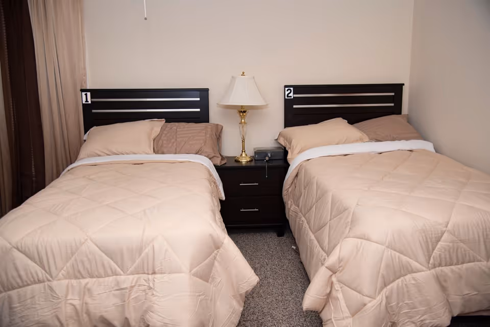 Two twin beds with beige comforters and numbered dark wood headboards separated by a nightstand and lamp.