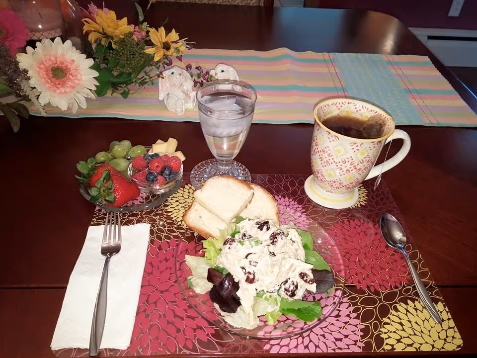 A dining table set with a colorful floral placemat holding a plate of chicken salad on lettuce with two slices of bread, a glass of iced water, a cup of tea with a tea bag, a small bowl of mixed berries, grapes, and cheese cubes. A fork and spoon are placed on either side of the placemat on a napkin and the table. In the background, there is a striped pastel table runner with artificial flowers and two small bunny figurines.