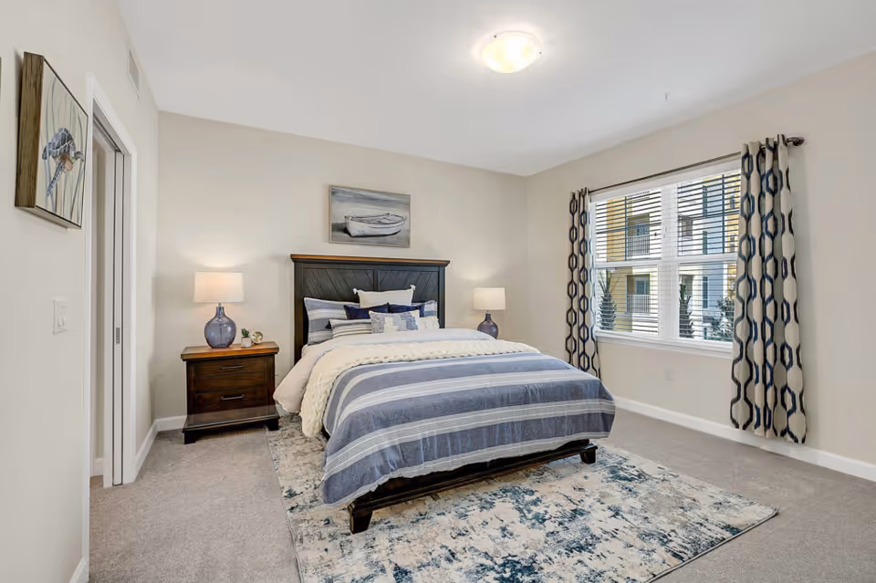 Well-lit bedroom with a bed dressed in blue-striped bedding, two nightstands with lamps, and a large window with patterned curtains.