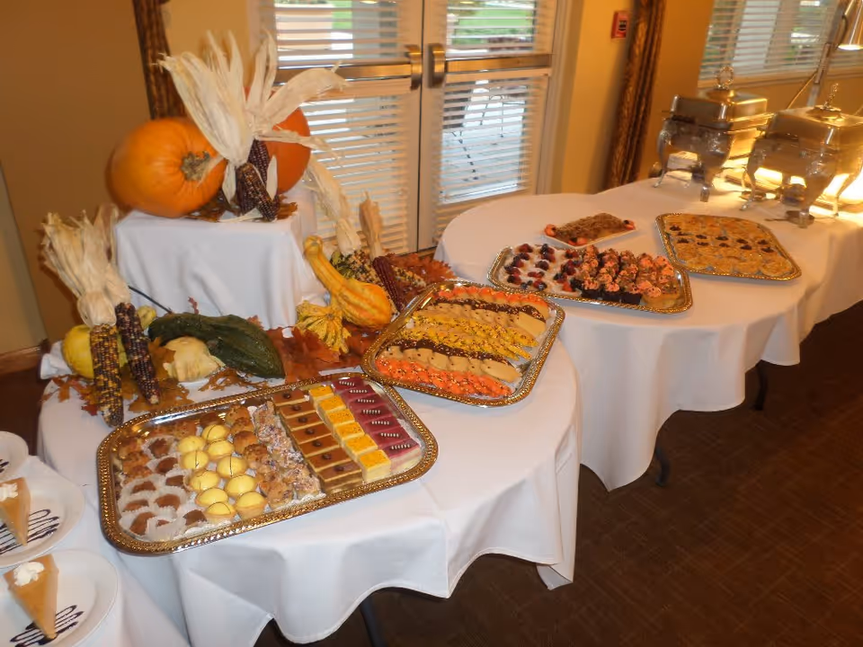 A buffet table set up with various desserts and appetizers on silver trays, decorated with pumpkins, gourds, and dried corn cobs. The table is covered with white tablecloths and is positioned near glass doors with blinds.